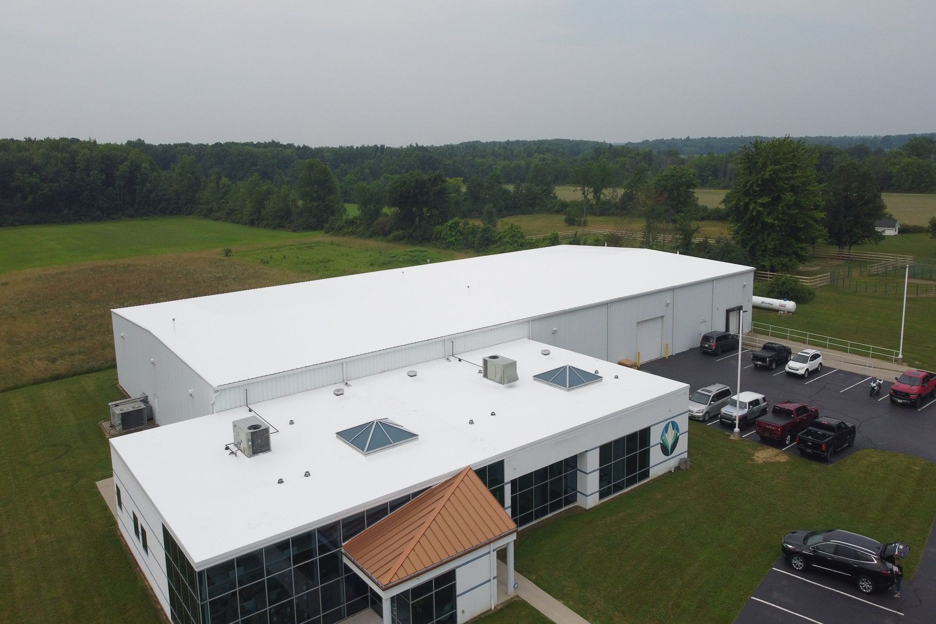 Aerial view of a white industrial building with a parking lot and surrounding green fields.
