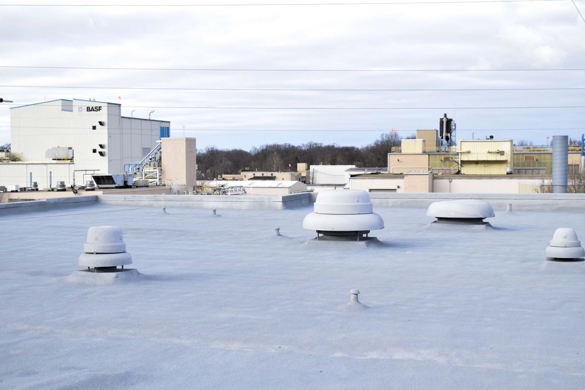 Snow-covered rooftop with several white vents and industrial buildings in the distance under a cloudy sky