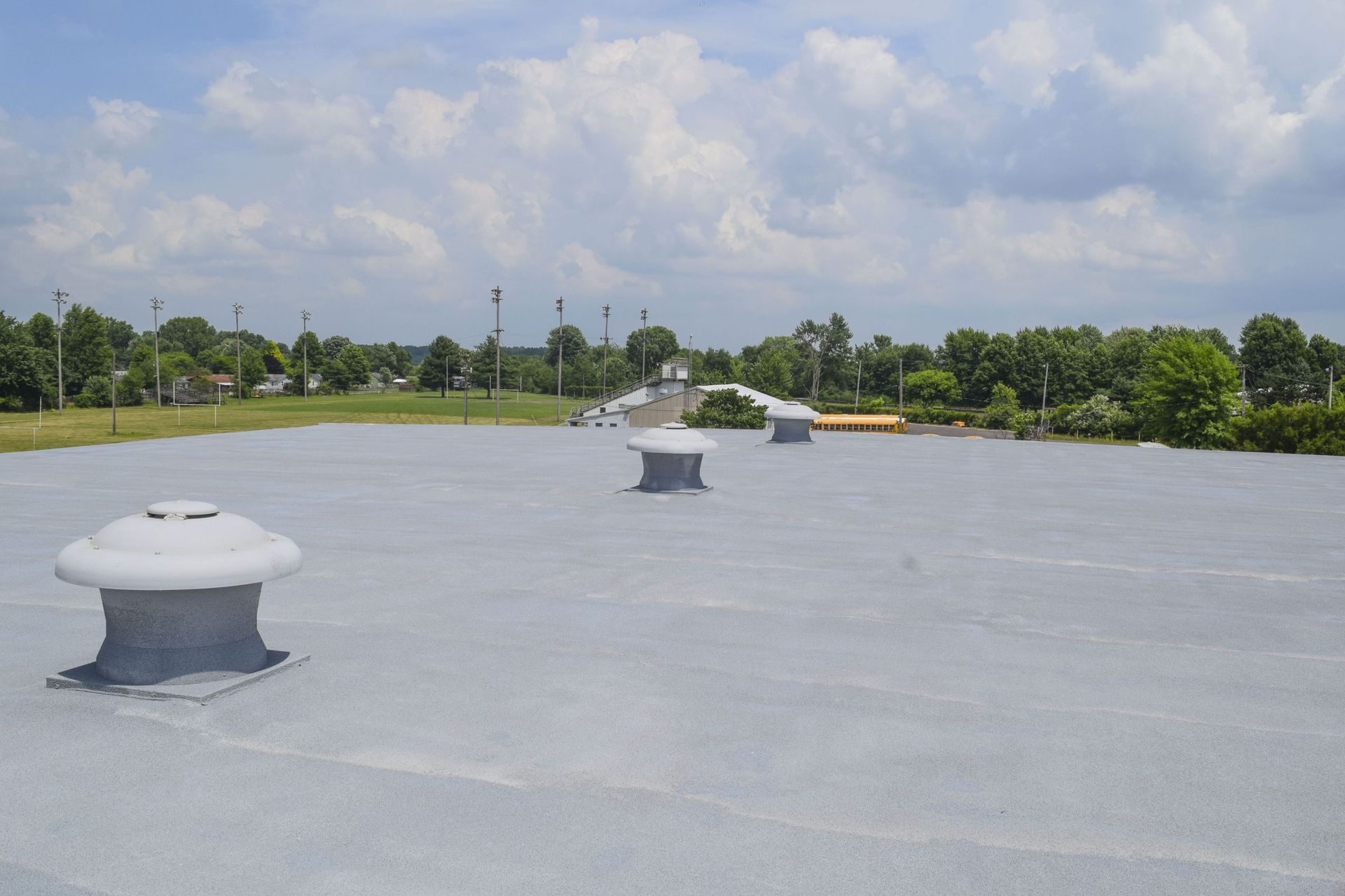 Flat gray rooftop with vents under a cloudy sky, bordered by trees in the distance.