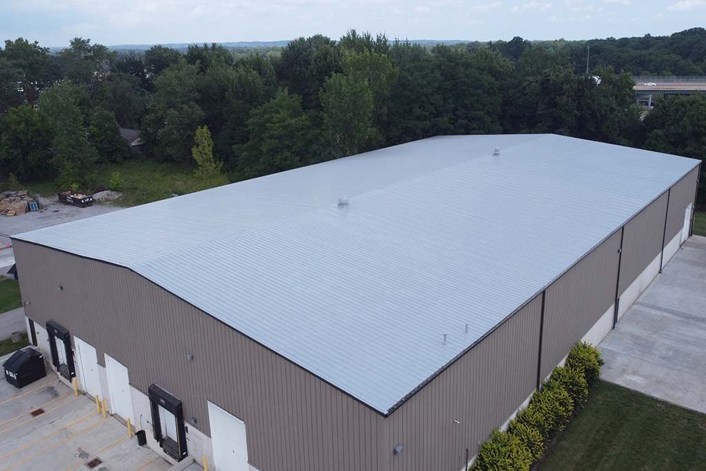 Aerial view of a large industrial warehouse with a light gray metal roof and tree-lined surroundings.