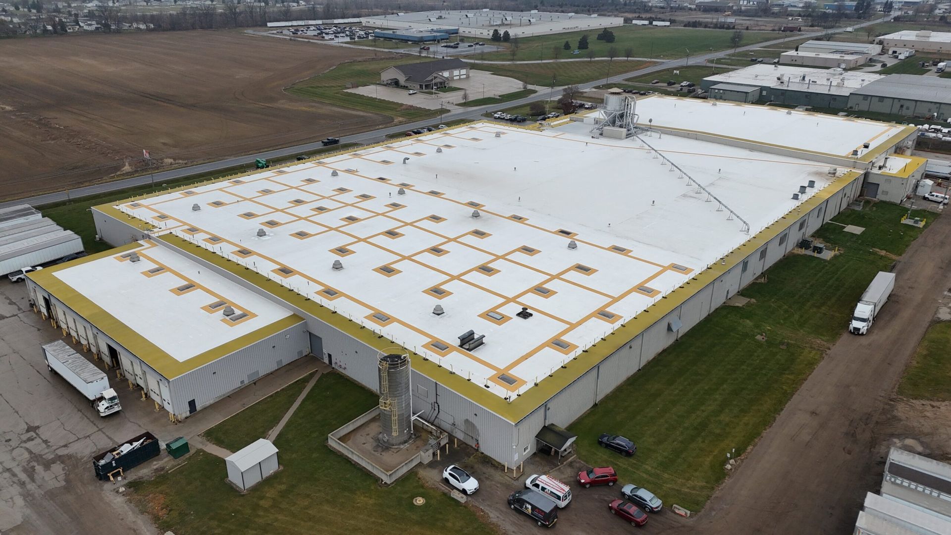 Aerial view of a large white-roofed warehouse with yellow trim in an industrial area and parked vehicles.