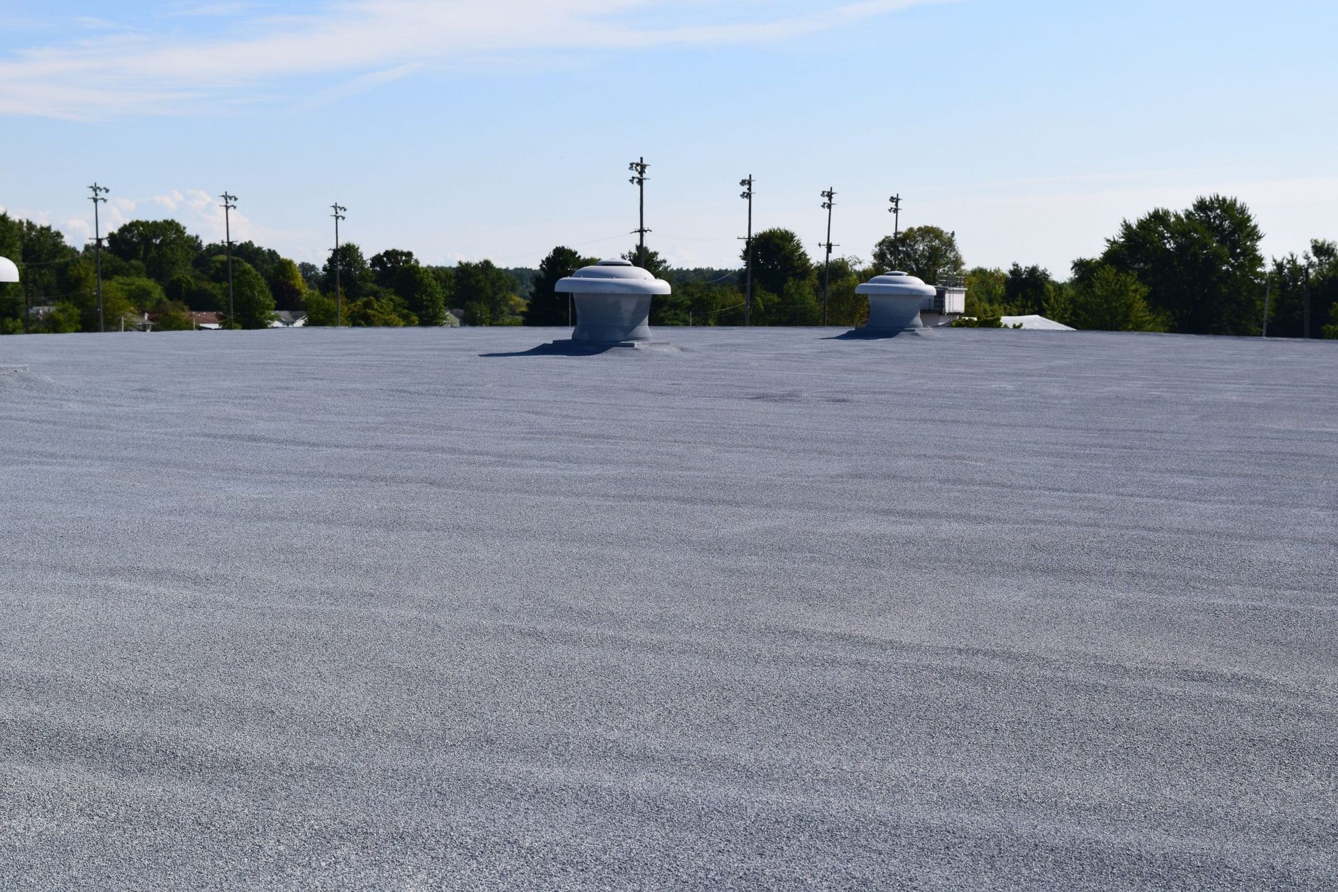 Flat gravel rooftop with two vents under a blue sky, trees and utility poles in the distance