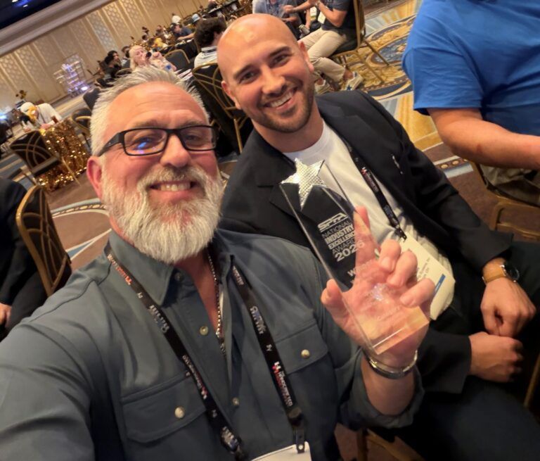 Two smiling men taking a selfie at a conference, with banquet tables and seated attendees behind them.