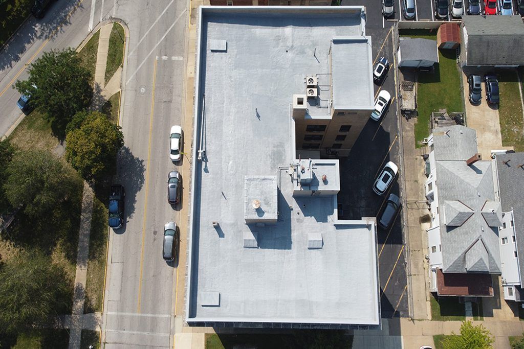 Aerial view of a flat-roofed building beside a road, with parked cars and nearby houses