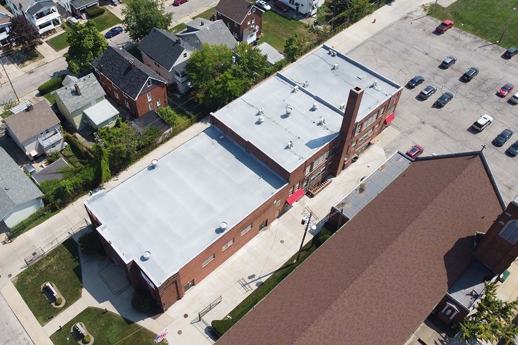 Aerial view of a red-brick industrial building with white roofs beside a parking lot and nearby houses.