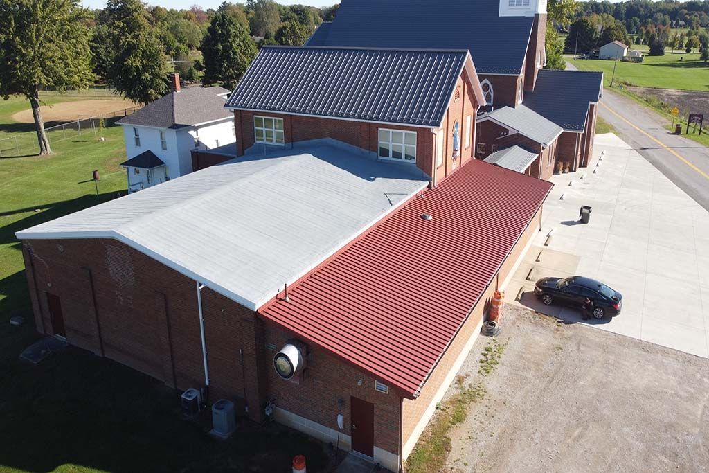 Aerial view of a brick building with red and gray roofs beside a gravel parking lot and road.
