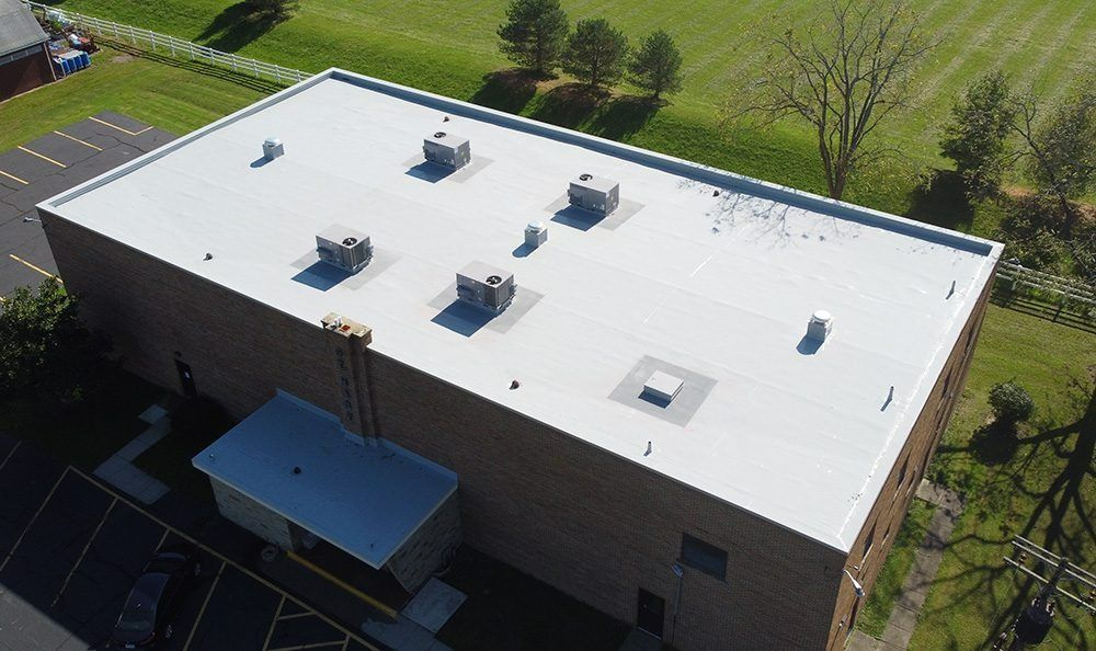 Aerial view of a flat white roof with vents and rooftop units beside a brick building and green lawn