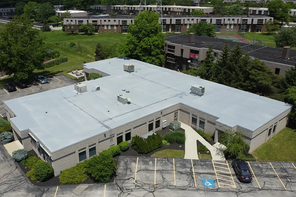 Aerial view of a white-roofed office building with parking lot, trees, and nearby buildings.