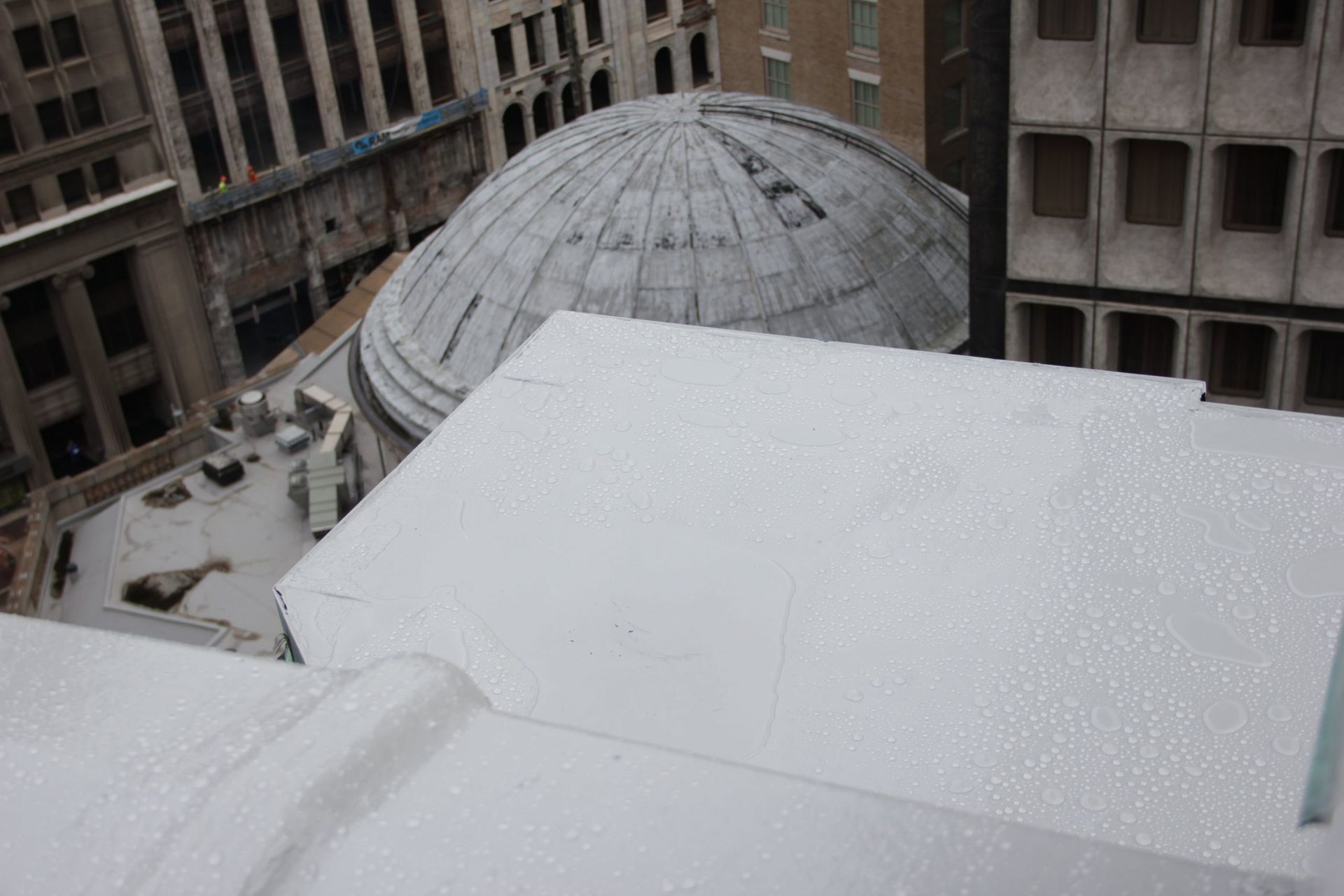 Snow-covered rooftop overlooking a city courtyard and domed building between tall office buildings