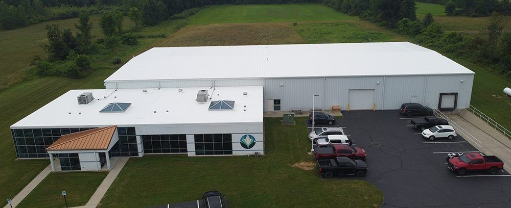 Aerial view of a white industrial building with parked cars and surrounding green fields.