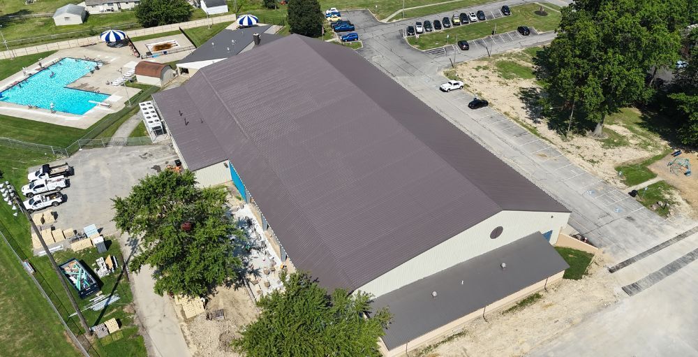 Aerial view of a large beige building with a dark roof beside a pool, parking lot, and trees.