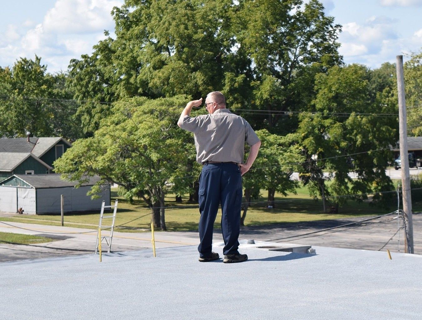 Man standing on a rooftop, shielding his eyes while looking into the distance near trees and houses.