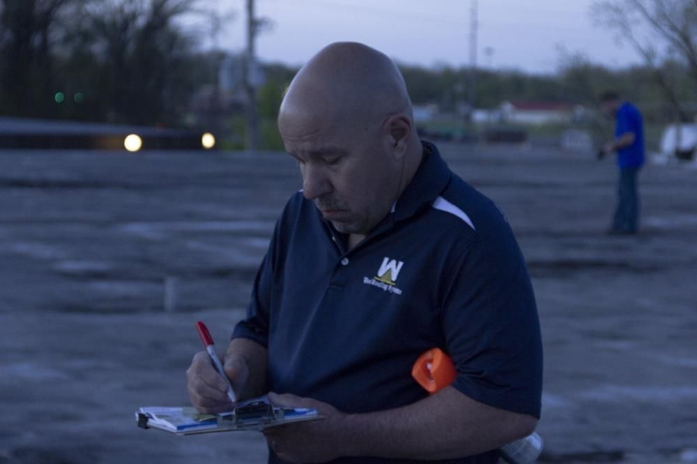 Man in navy shirt writing on a clipboard outdoors at dusk, with blurred lights and a person in blue behind him