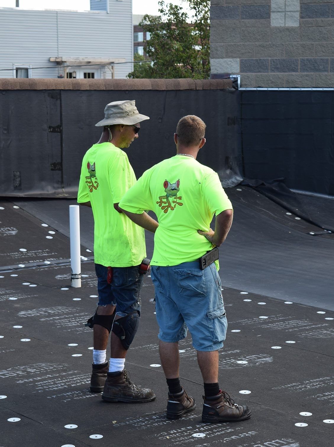 Two workers in neon green shirts stand on a black rooftop, looking over the area.