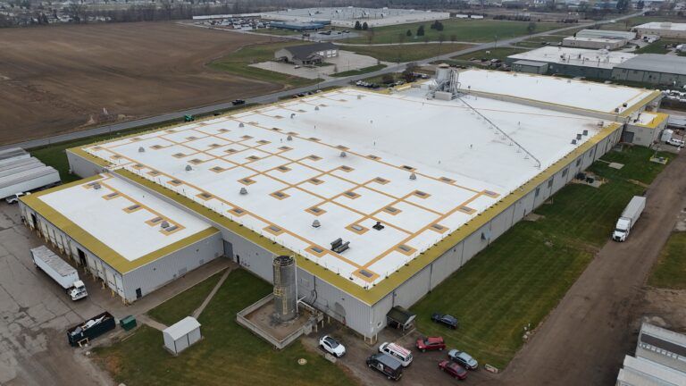 Aerial view of a large industrial building with a white roof and yellow trim in a rural area.