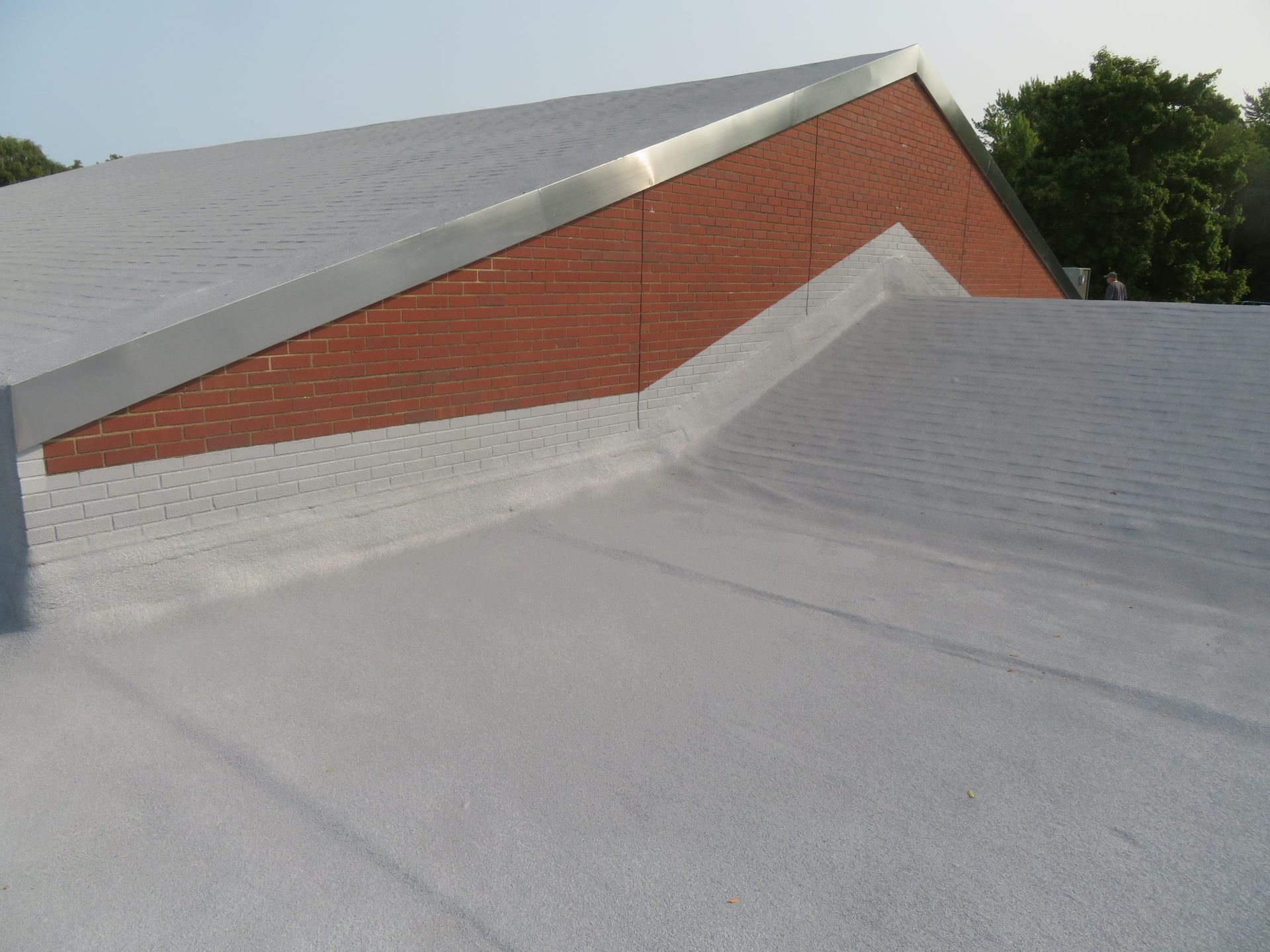 Flat gray rooftop with a red brick wall and silver metal trim against trees and sky