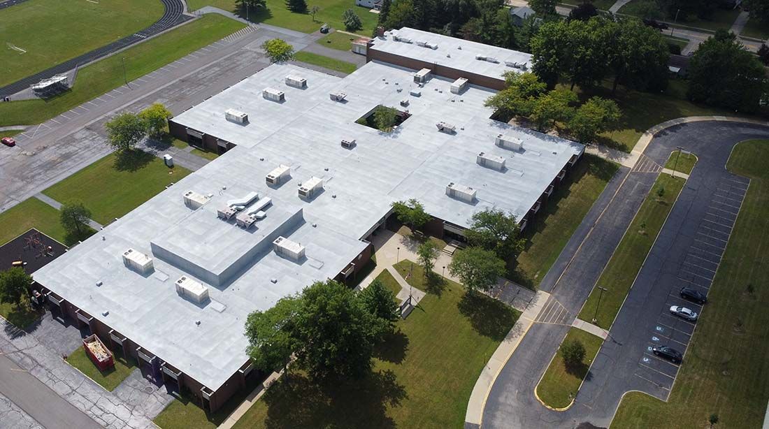 Aerial view of a large white flat-roofed building beside a parking lot and road, surrounded by trees.