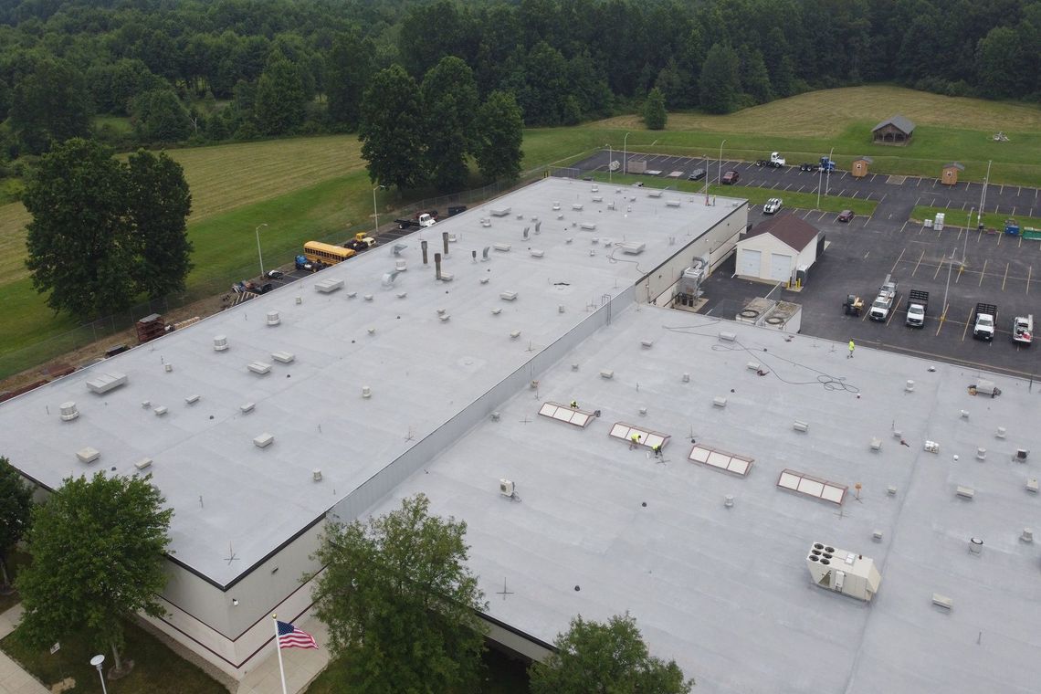 Aerial view of a large gray-roofed building beside a parking lot and trees.