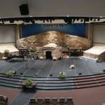 Empty church stage with blue carpet, stone backdrop, and potted plants in front of seating.