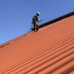 Worker on a red roof using a safety harness and tool, beneath a clear blue sky