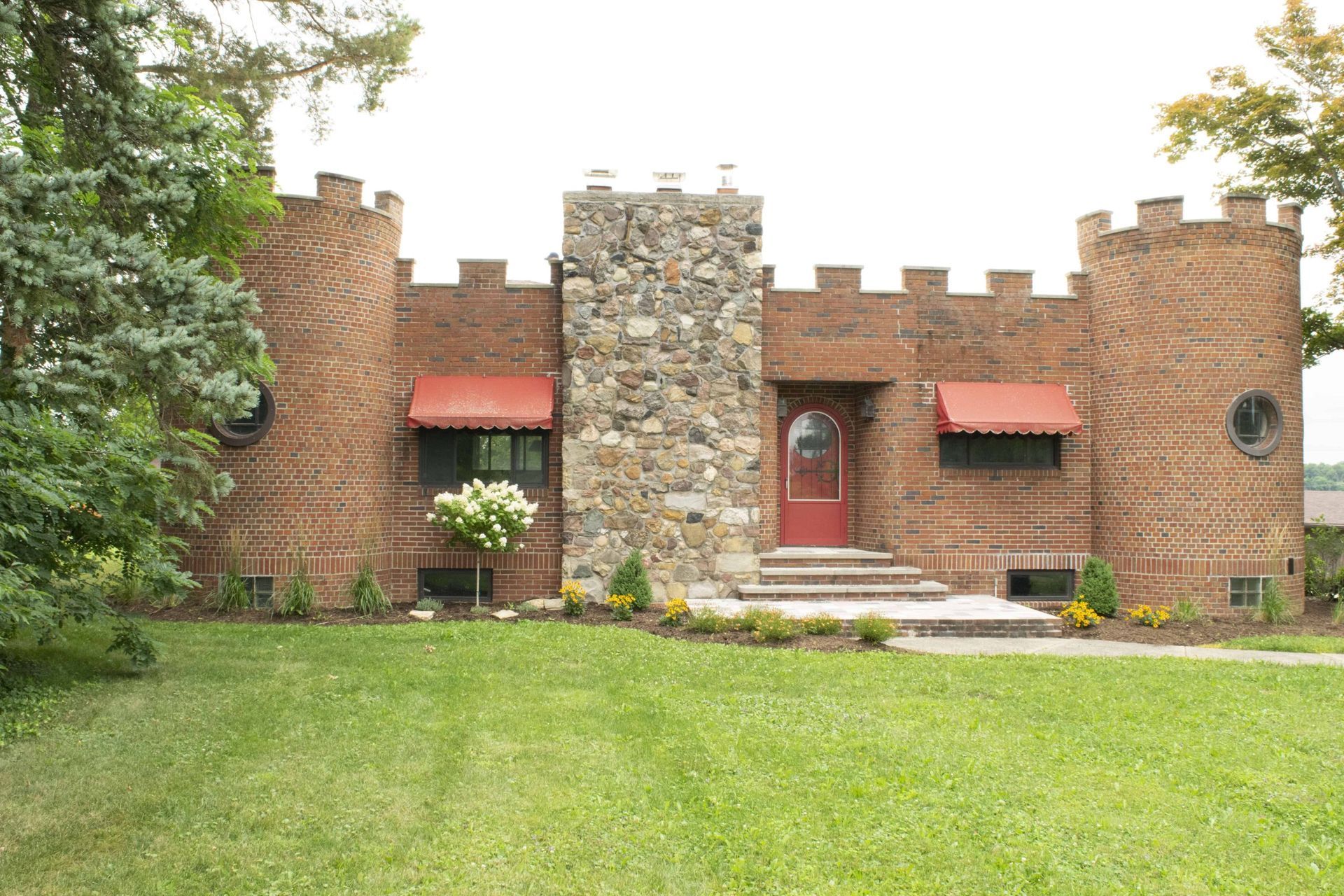 Brick castle-like house with red awnings, central red door, and a large grassy lawn