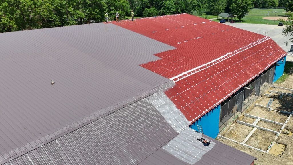 Aerial view of a building with gray, red, and blue metal roofs next to a wooded area.