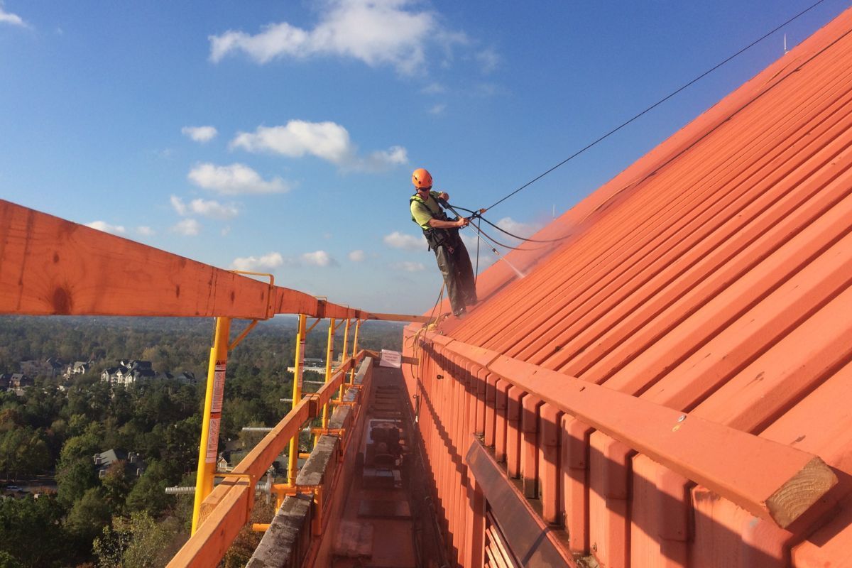 Worker in safety gear on an orange rooftop, balancing beside a steep sloped roof under a blue sky.