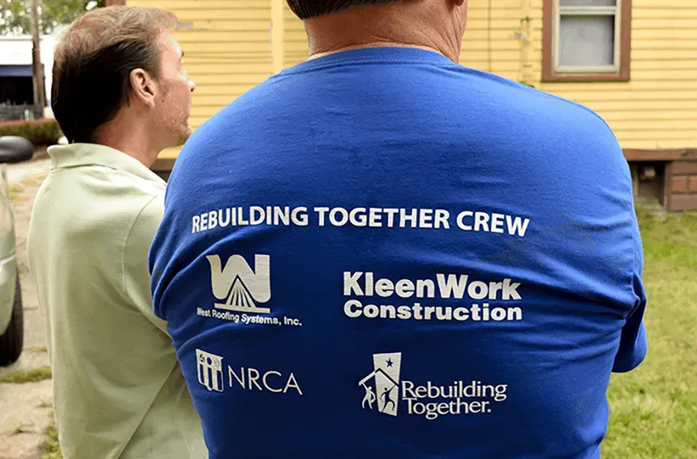 Two volunteers in branded shirts outdoors, with “Rebuilding Together Crew” and KleenWork Construction logos visible.