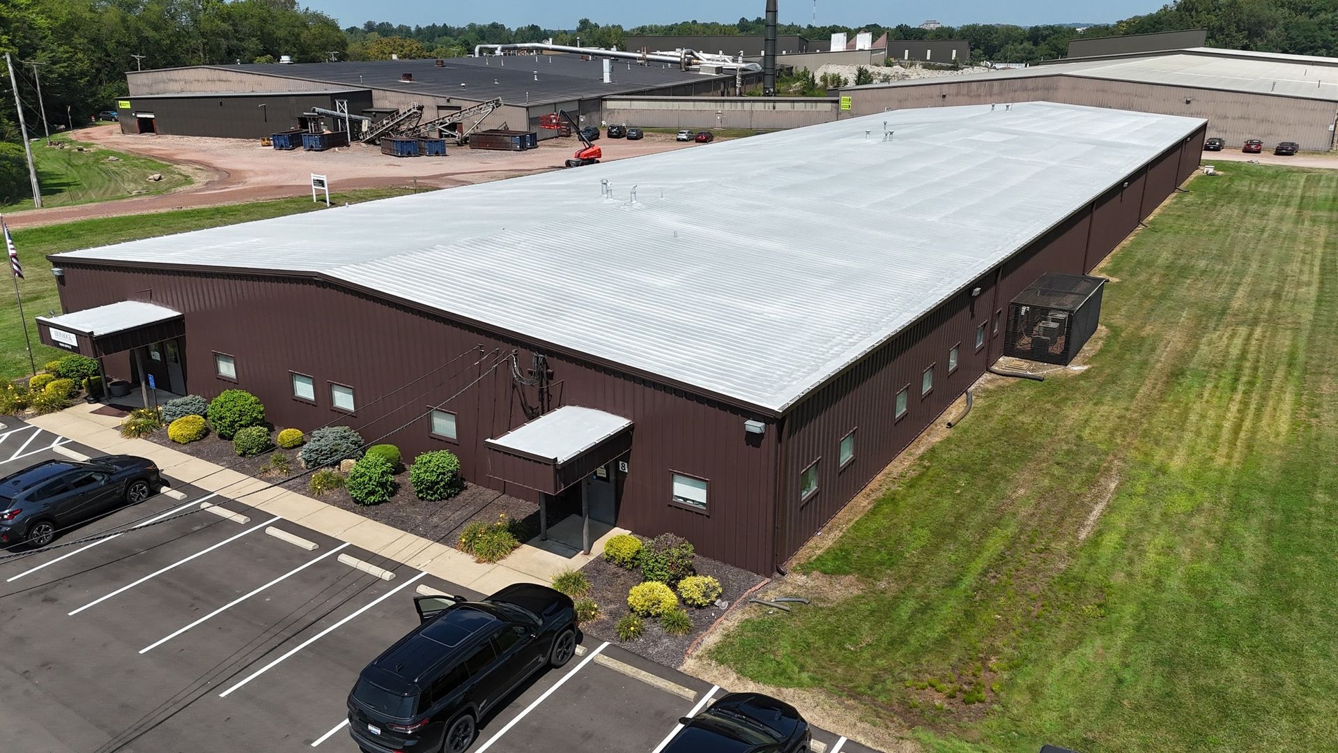 Aerial view of a long brown industrial building with a white roof and parking lot beside it