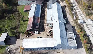 Aerial view of long white-roofed buildings beside a road and green trees.