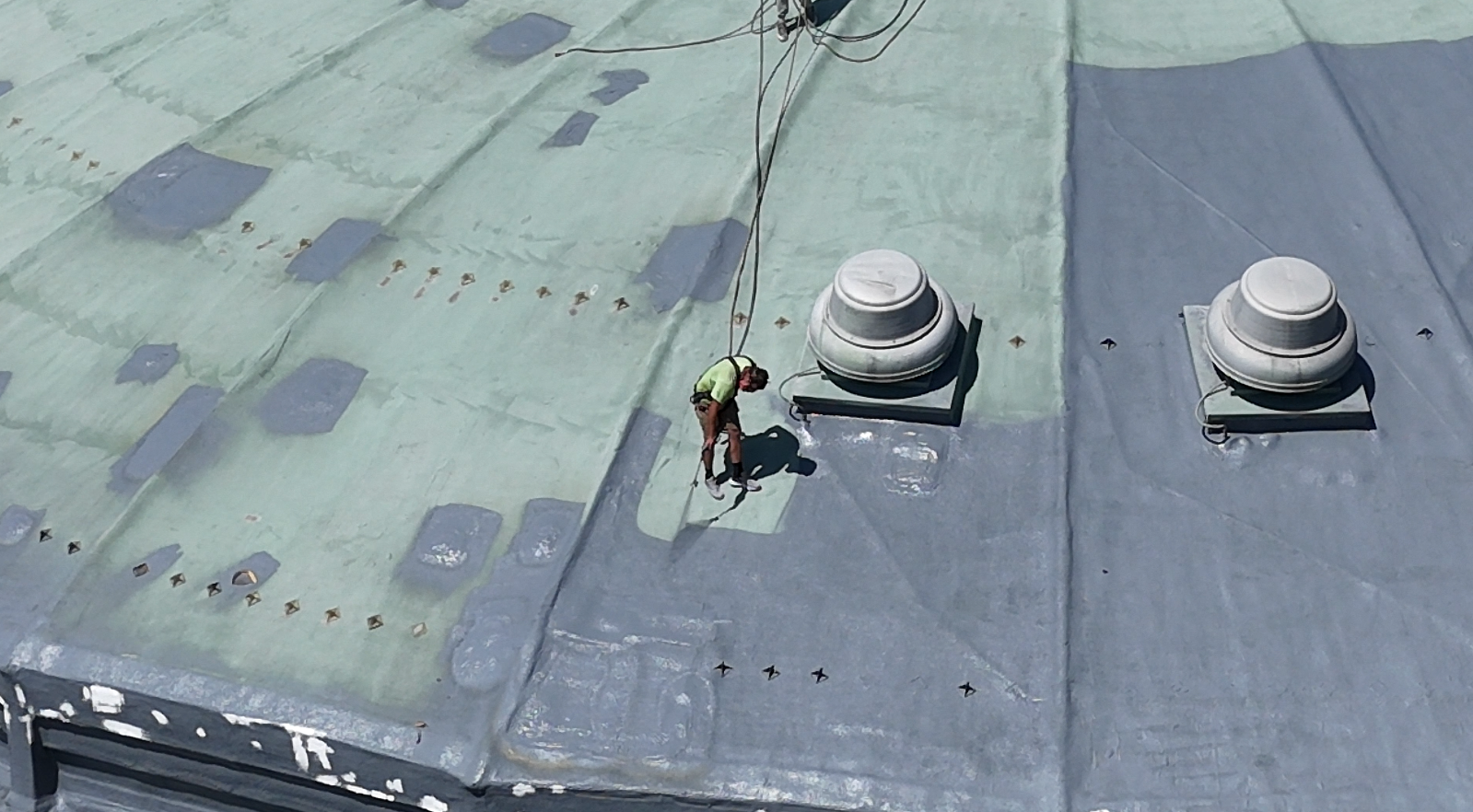 Two workers on a large green metal roof near two vent domes, viewed from above
