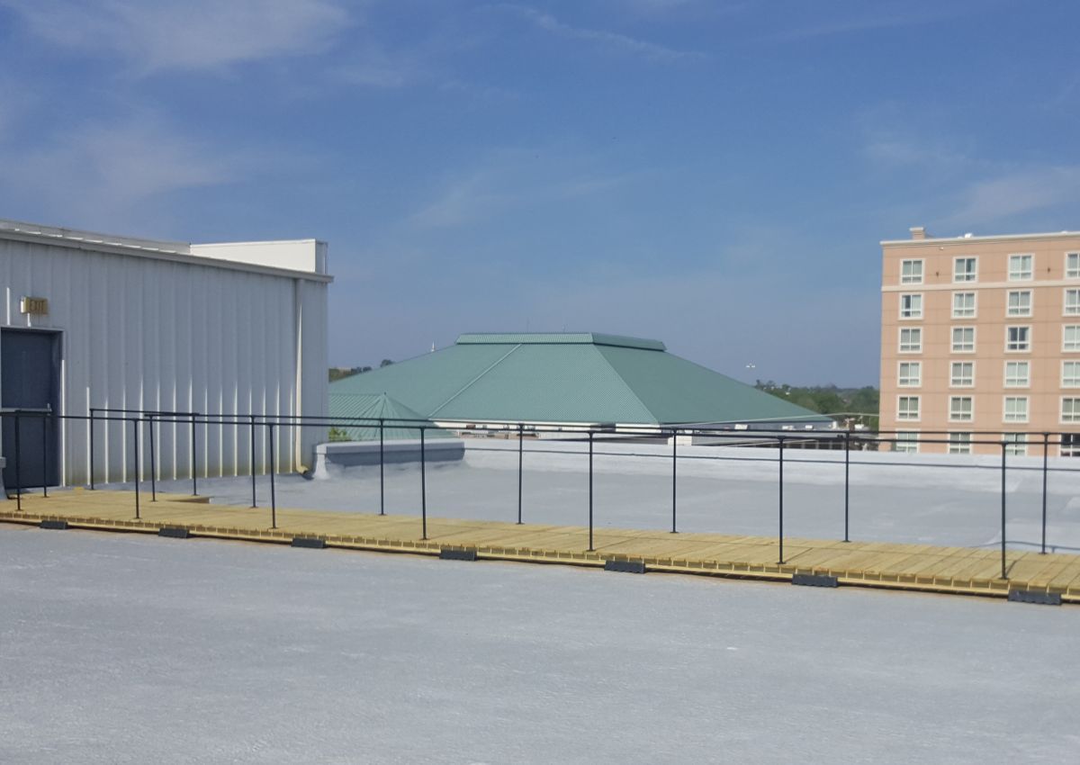 Rooftop fenced area with a green structure, next to a white building and brick apartment building under a blue sky