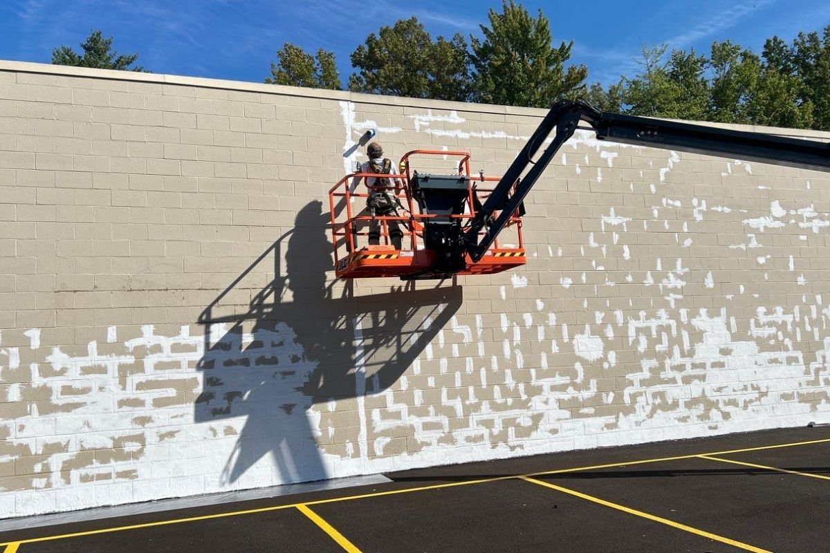 Worker in orange lift painting a tan wall with white patches in a parking lot.