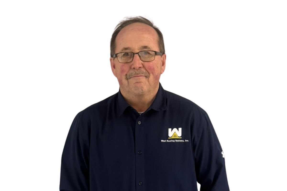 Man in navy shirt with logo, standing against a white background, wearing glasses and smiling gently