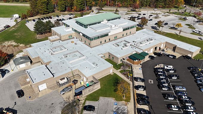 Aerial view of a large beige building with a green roof and parking lot surrounded by trees