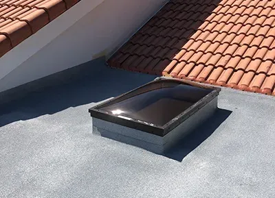 Rooftop skylight with a dark frame on a gray flat roof beside red tiled roofing