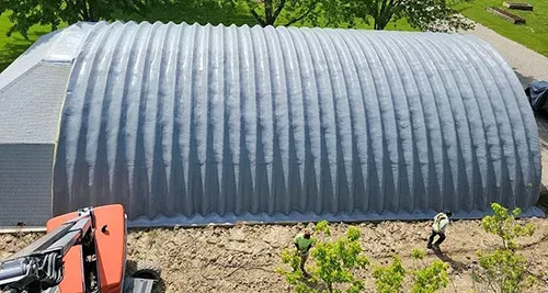 Gray corrugated metal Quonset hut beside a driveway with a small excavator in the foreground