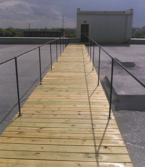 Wooden rooftop walkway with black railings leading to a small gray utility building.