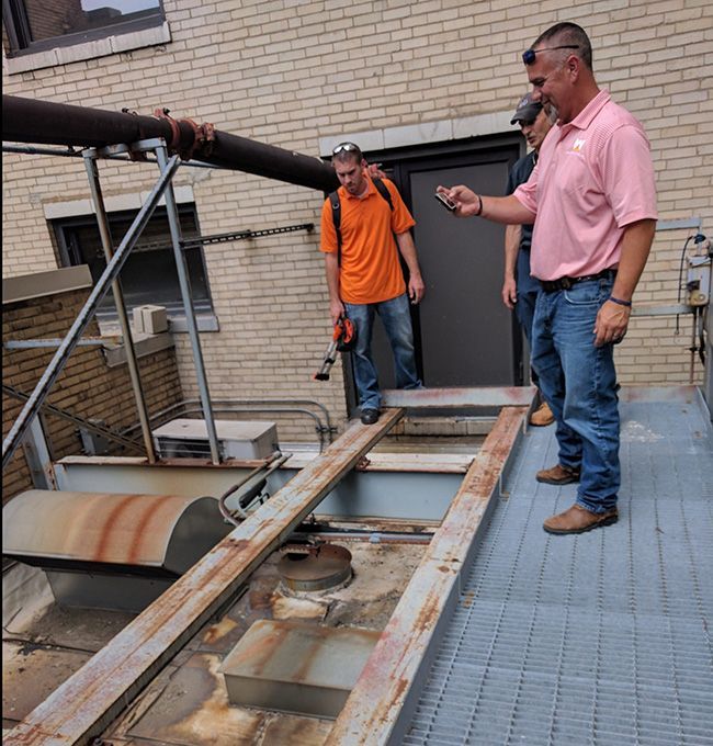 Two workers inspecting a rusted industrial tank beside a brick building, one holding a phone.