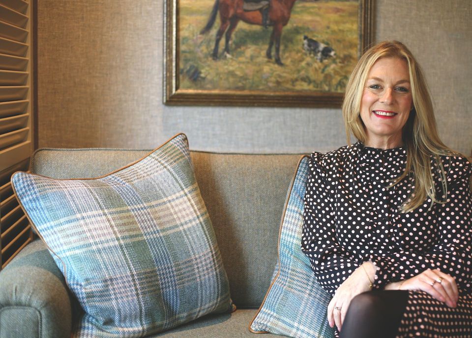 Woman sitting on a couch smiling, wearing polka dot top. Painting of a horse and dog behind her.