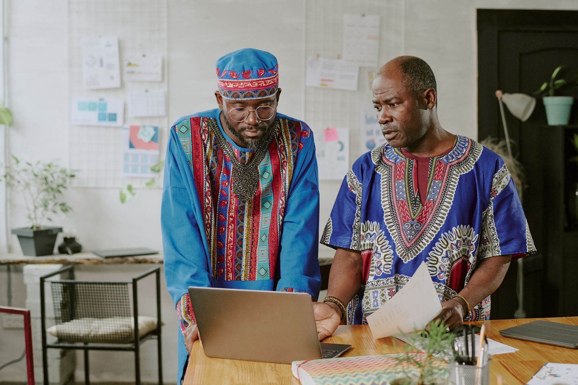 Two men in colorful African attire look at a laptop, one holding papers, in a brightly lit office setting.