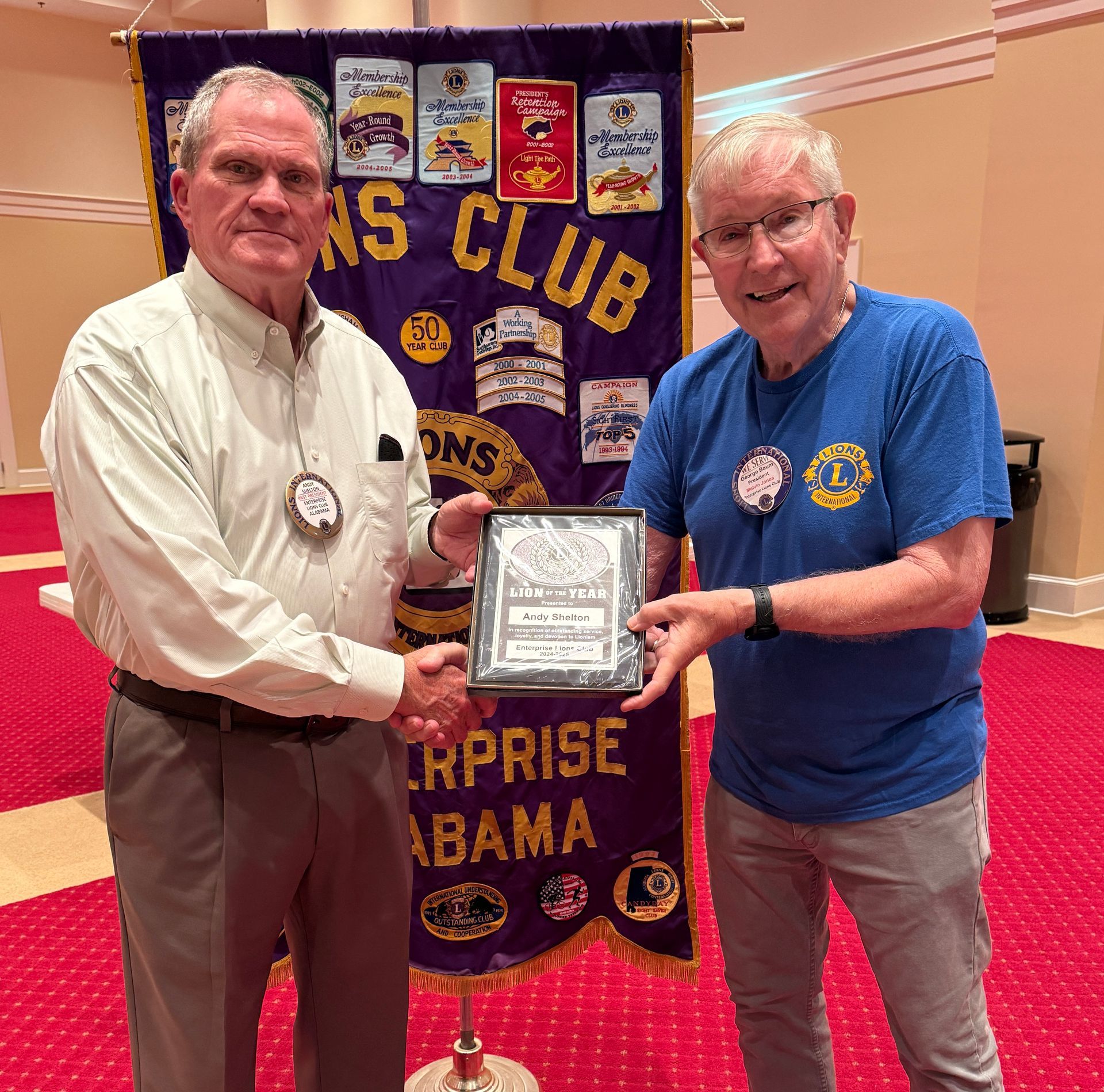 Two men shake hands, one presents a plaque near a Lions Club banner.