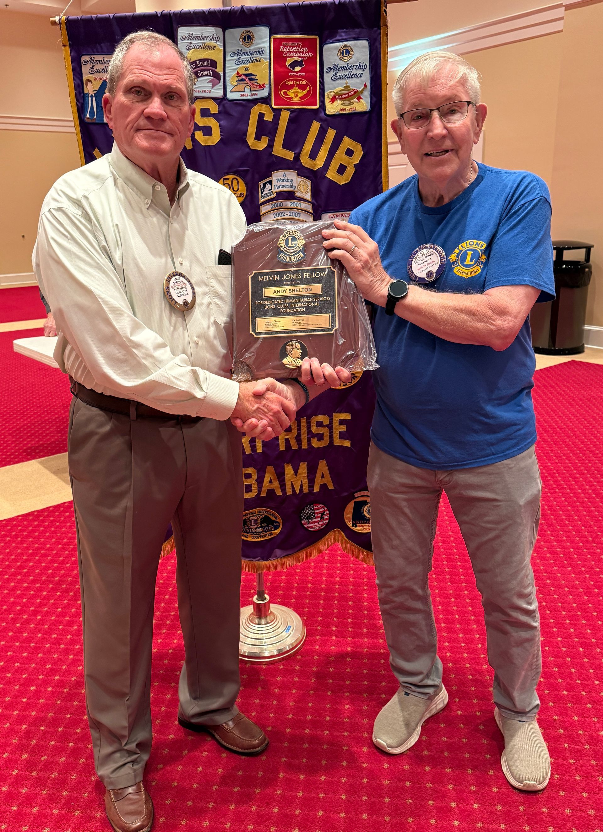 Two men shaking hands, one presenting an award in front of a Lions Club banner.