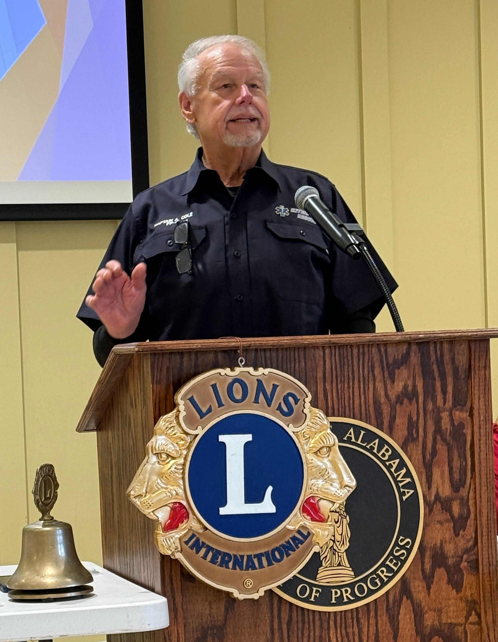 Man speaking at a podium with Lions Club emblem, microphone, and a small bell on the podium