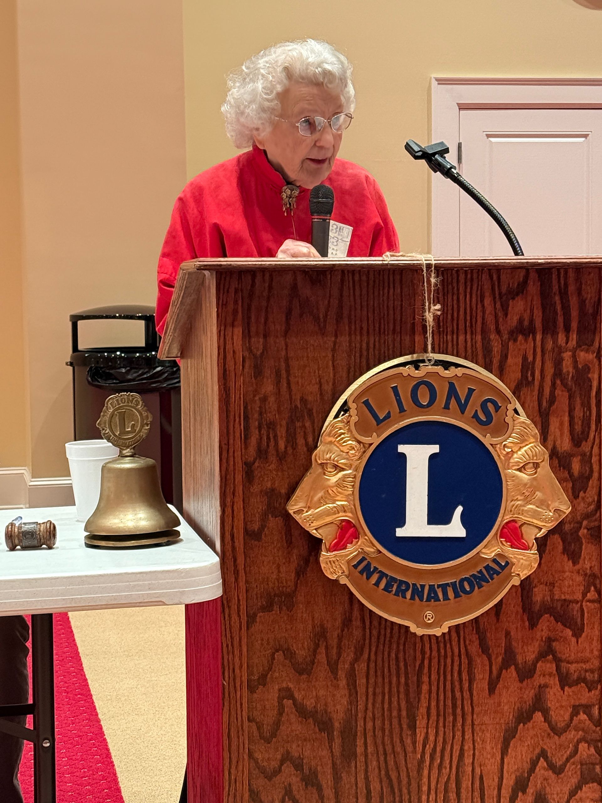 Woman speaking at a Lions Club podium. She wears a red shirt and glasses; the podium displays a Lions logo.