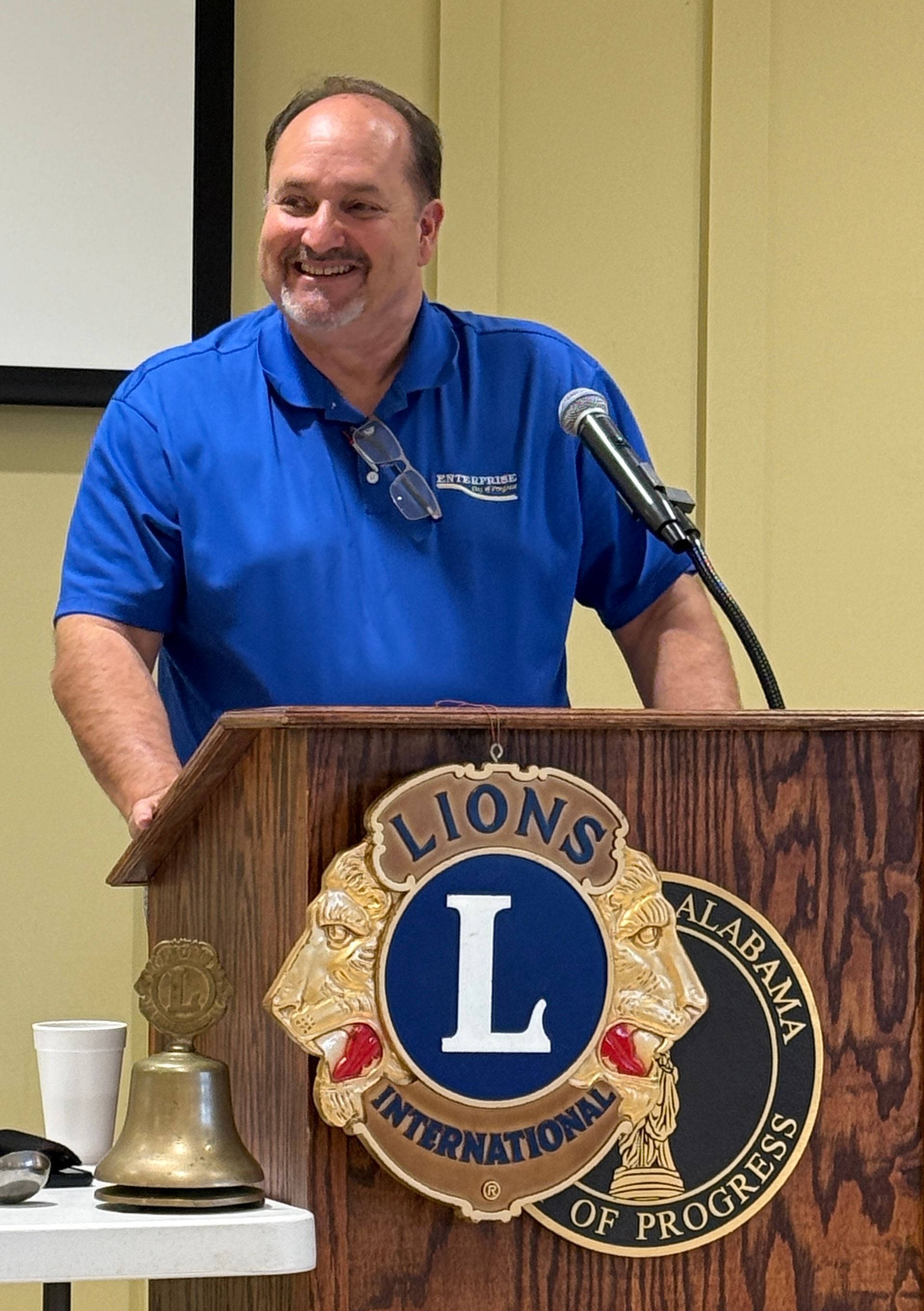 Man speaking at a Lions Club podium with a microphone, wearing a blue shirt and smiling.