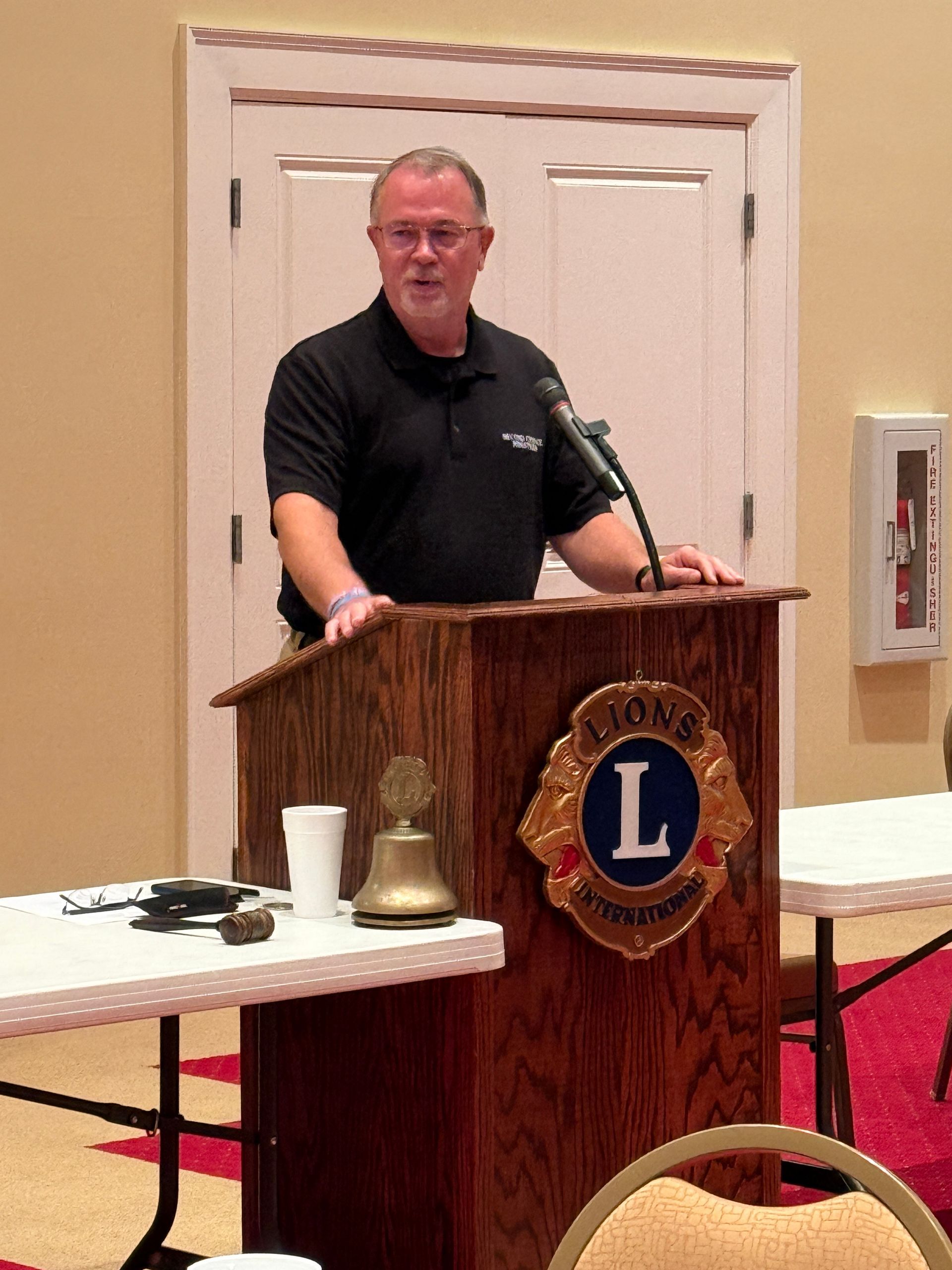 Man speaking from a Lions Club podium. He wears a black shirt, stands at a wooden lectern with Lions Club emblem.