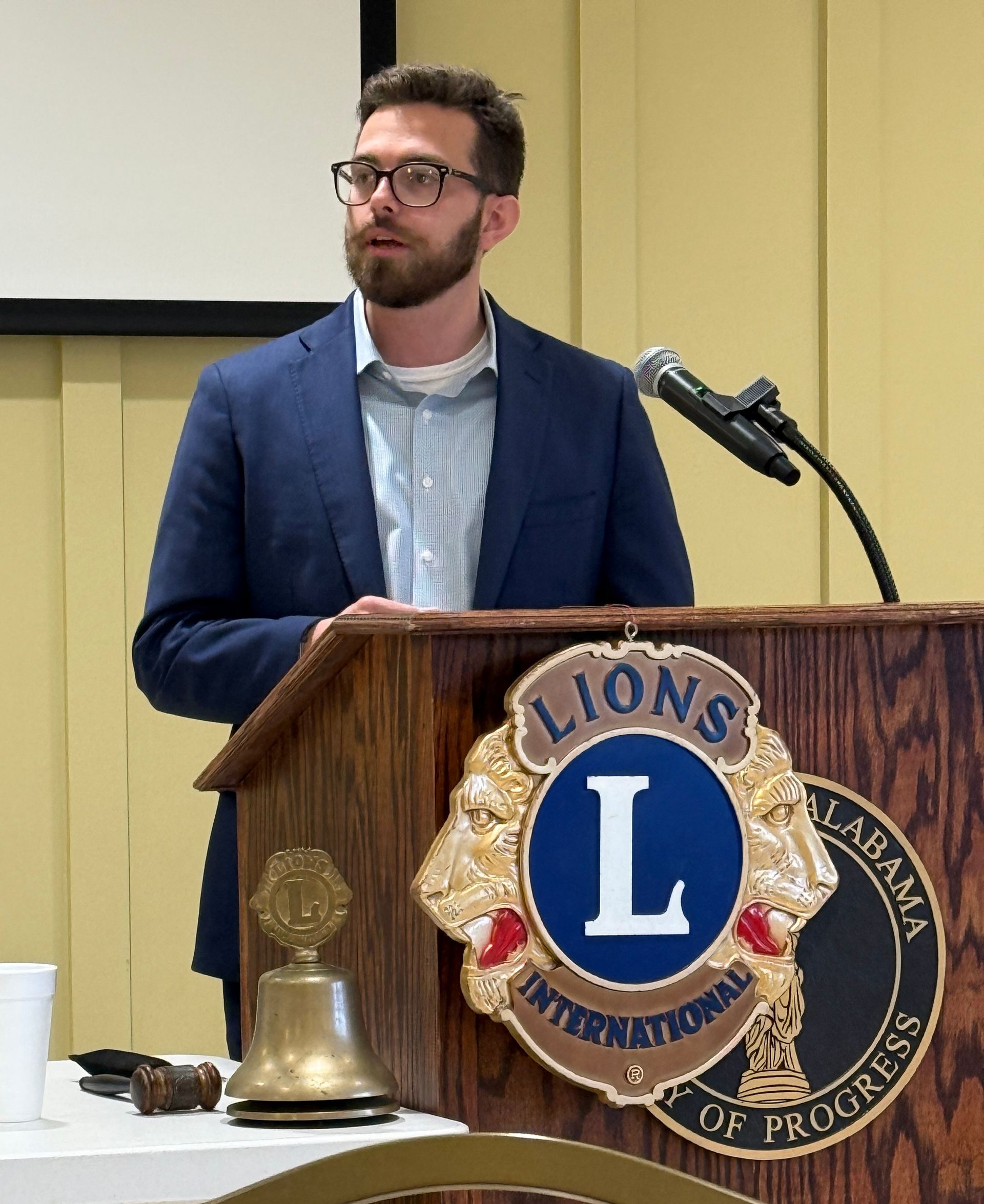 Man speaking at a Lions Club podium, wearing glasses and blue blazer. Lions Club logo and gavel visible.
