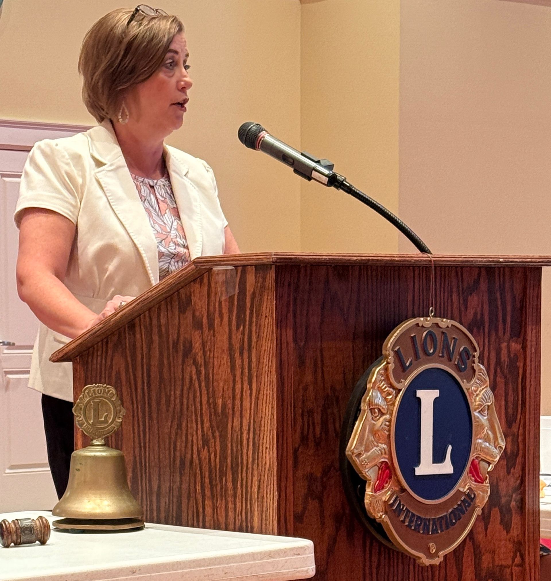 Woman speaking at a podium with Lions Club emblem; cream blazer, microphone, bell, and gavel.