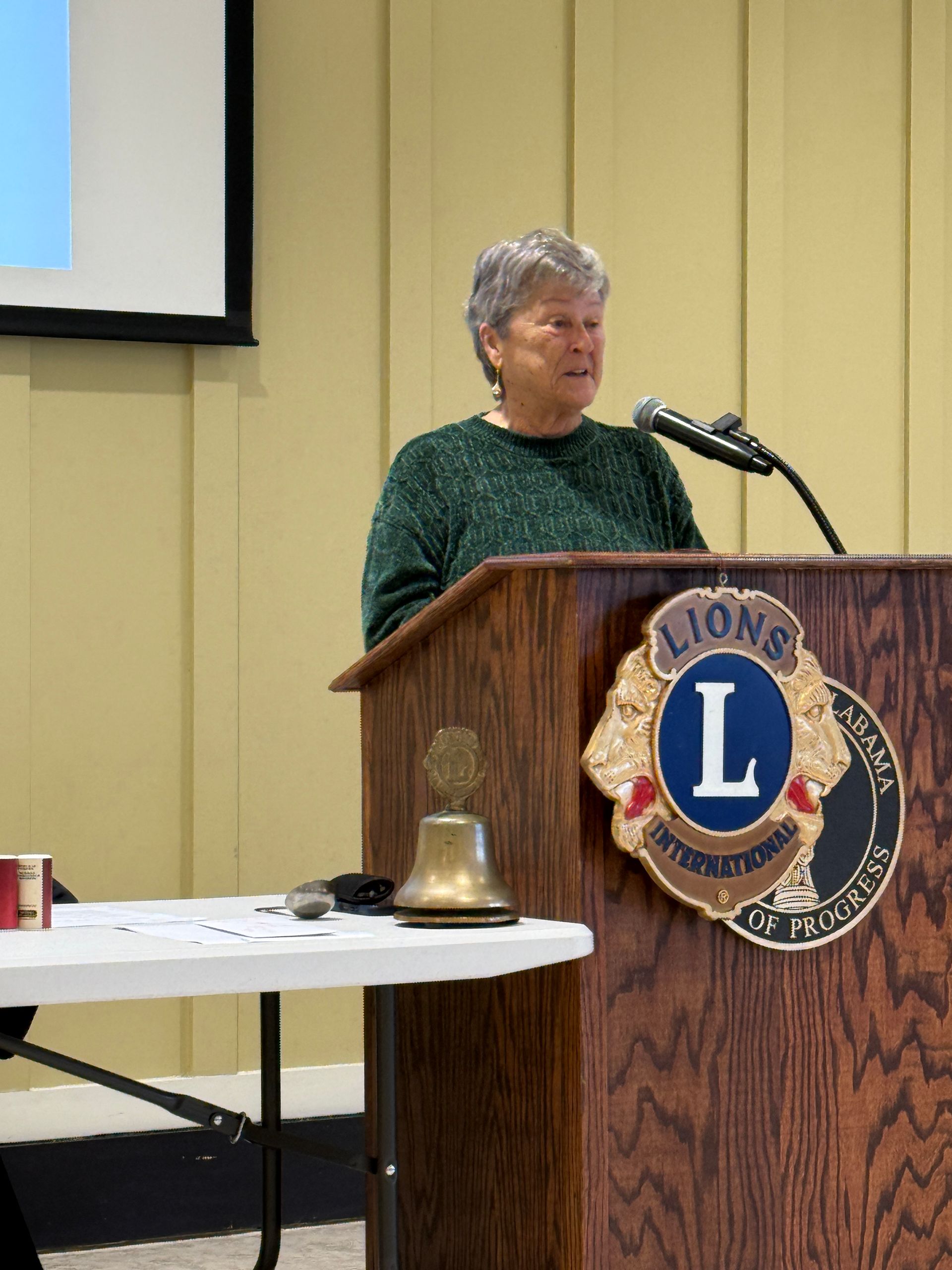Woman speaking at a Lions Club podium beside a microphone in a meeting hall.